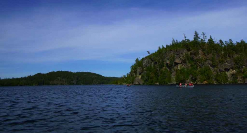 a canoe is paddled on a calm blue lake along side a tree-lined shore 